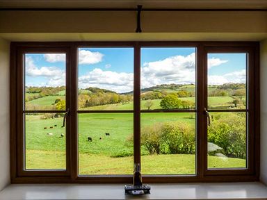 A view of green fields with grazing cows and rolling hills through a wooden framed window at Glebe Farm Cottage in Llandegveth near Cwmbran