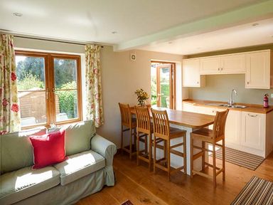 A living area with a green sofa and red cushion next to a wooden dining table with four chairs and a kitchen in the background at Glebe Farm Cottage in Llandegveth near Cwmbran