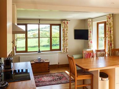 A kitchen and dining area with wooden table and chairs windows with floral curtains and a tv on the wall at Glebe Farm Cottage in Llandegveth near Cwmbran