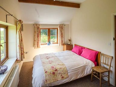 A bedroom with a bed with pink pillows next to a wooden chair and windows with floral curtains at Glebe Farm Cottage in Llandegveth near Cwmbran