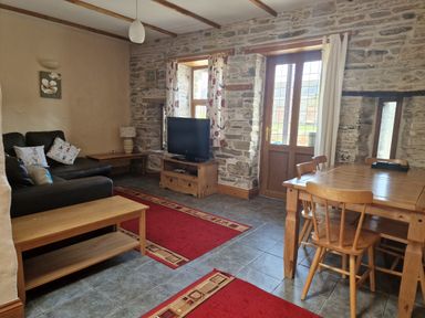 A living room with a black sofa, wooden coffee table, TV on a wooden stand, dining table with chairs, stone walls, and red rugs at Kingfisher Cottage in Llanboidy