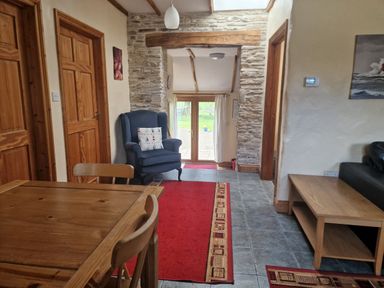A hallway with a wooden dining table and chairs a blue armchair with a cushion red rugs on a tiled floor wooden doors a stone archway and glass doors leading outside at Kingfisher Cottage in Llanboidy