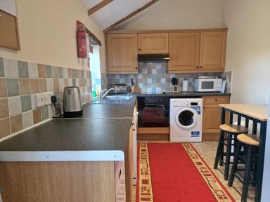 A kitchen with wooden cabinets appliances a red rug and a small dining table with stools at Kingfisher Cottage in Llanboidy