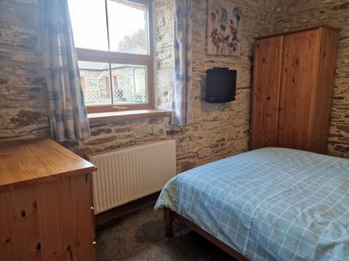 A bedroom with a bed covered in blue checkered bedding a wooden wardrobe a wooden dresser a window with curtains and a wall-mounted TV at Kingfisher Cottage in Llanboidy