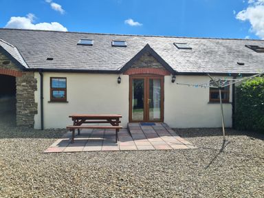 An outdoor patio area with a wooden picnic table and a rotary clothesline in front of a building with a tiled roof and two small windows at Kingfisher Cottage in Llanboidy