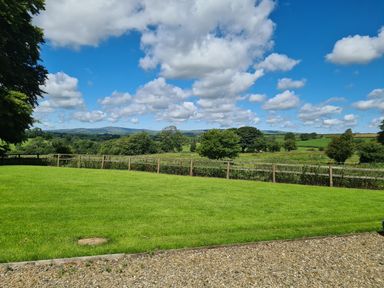 A grassy yard with gravel path and wooden fence overlooking fields and trees under a cloudy blue sky at Kingfisher Cottage in Llanboidy