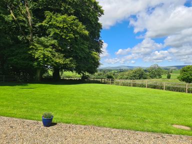 A grassy yard with a small blue flower pot near a gravel area with trees and hills in the background at Kingfisher Cottage in Llanboidy