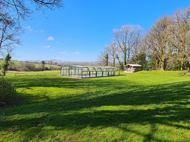 A fenced pool with a glass cover and a small wooden cabin in a green field with trees at Kingfisher Cottage in Llanboidy