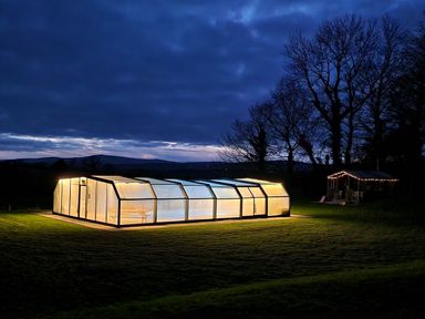 A lit greenhouse on grass with a small building decorated with string lights at Kingfisher Cottage in Llanboidy
