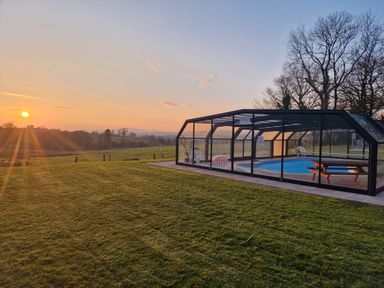 An outdoor enclosed swimming pool with chairs and picnic table on a grassy field at sunset at Kingfisher Cottage in Llanboidy