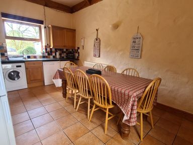 A kitchen with a table covered by a red checkered tablecloth surrounded by wooden chairs a washing machine dishwasher wooden cabinets and tiled floor at Swallow Cottage in Llanboidy