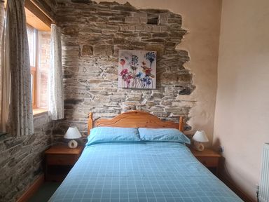 A bedroom with a stone wall behind a wooden bed with blue bedding and pillows two nightstands with white lamps and a window with curtains at Swallow Cottage in Llanboidy