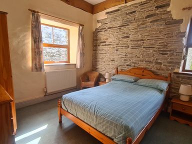 A bedroom with a wooden bed with blue bedding a stone wall two windows curtains a wicker chair and bedside tables with lamps at Swallow Cottage in Llanboidy