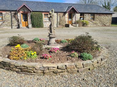 A circular stone flower bed with a statue in the center and a stone building in the background at Swallow Cottage in Llanboidy
