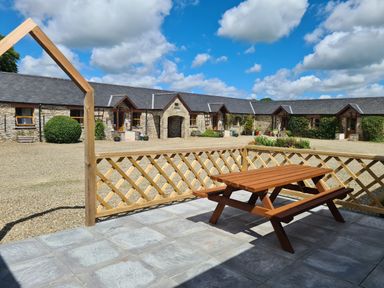 An outdoor stone patio with a wooden picnic table and lattice fence facing a stone building with multiple doors and windows at Swallow Cottage in Llanboidy