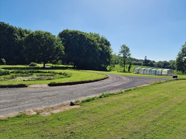 A gravel driveway curving around a pond with trees and a covered swimming pool in a grassy landscape at Swallow Cottage in Llanboidy
