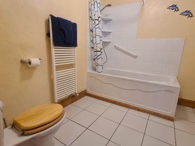 A bathroom with a toilet with wooden seat a white towel rack with blue towels and a bathtub with a shower curtain and dolphin wall decorations at Sandpiper Cottage in Llanboidy