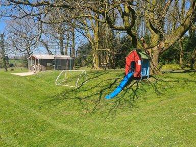 A playground with a small soccer goal a red playhouse with a blue slide a wooden swing set and a wooden cabin among trees at Sandpiper Cottage in Llanboidy