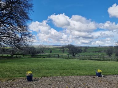 A lawn with two flower pots in the foreground and fields with trees under a cloudy sky at Sandpiper Cottage in Llanboidy