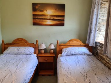 A bedroom with two single wooden beds a nightstand with two lamps a sunset painting and a window with curtains at Puffin Cottage in Llanboidy