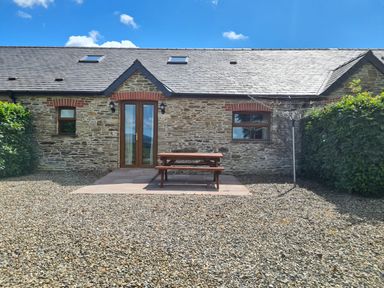 A stone house with wooden doors and windows a wooden picnic table on a paved area and gravel ground at Puffin Cottage in Llanboidy