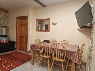 A dining area with a table covered with a checkered cloth and six wooden chairs with a wall-mounted TV and a wooden door at Puffin Cottage in Llanboidy