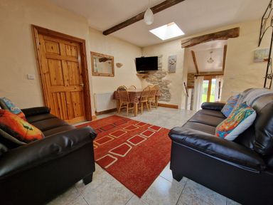 A sitting room with two black sofas a red rug a wooden dining table with chairs and a wall mounted TV at Puffin Cottage in Llanboidy