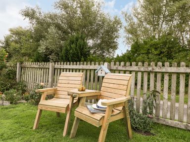 Two wooden chairs with a small table between them on a lawn in a garden with a wooden fence and trees at Pear Tree Cottage Mark near Burnham-on-Sea