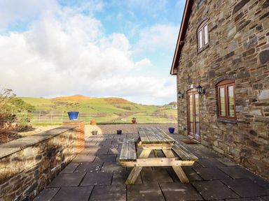 An outdoor patio with a wooden picnic table next to a stone house with hills in the background at The Old House in Llanidloes