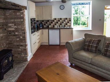 A small kitchen area with stove and cabinets next to a stone fireplace and living room with sofa and cushions at Cotswold View Clifford Chambers near Stratford-upon-Avon