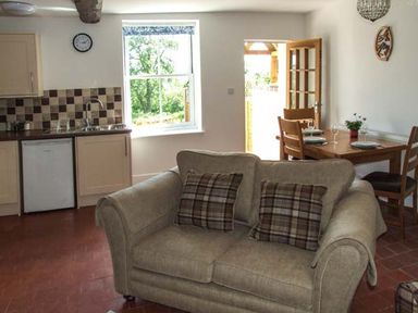 A kitchen and dining area with a beige sofa in the foreground a dining table set with plates and glasses and a window showing greenery at Cotswold View in Clifford Chambers near Stratford-upon-Avon