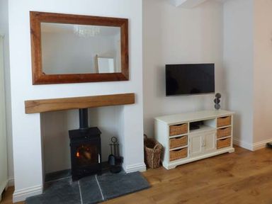 A room with a wood burning stove under a wooden mantel and a wall mounted TV above a cabinet with wicker baskets at Silversands Cottage in Amble-by-the-Sea