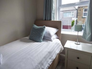 A bedroom with a single bed next to a window and a bedside table with a lamp and flowers at Silversands Cottage in Amble-by-the-Sea