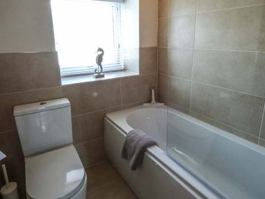 A bathroom with a toilet, bathtub with glass screen, towel on tub, and a window with a seahorse ornament at Silversands Cottage in Amble-by-the-Sea