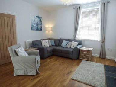 A living room with a gray corner sofa a patterned armchair wooden floor and a window with curtains at Silversands Cottage in Amble-by-the-Sea
