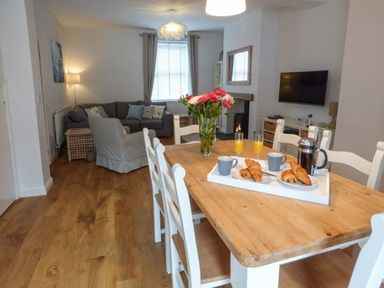 A dining area with a wooden table set with breakfast items and a vase of flowers and a living room with a sofa and armchair at Silversands Cottage in Amble-by-the-Sea
