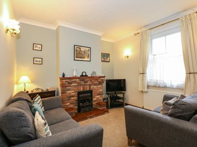 A living room with two gray sofas a brick fireplace a small television on a stand and a window with curtains at 5 Melinda Cottages in East Runton near Cromer