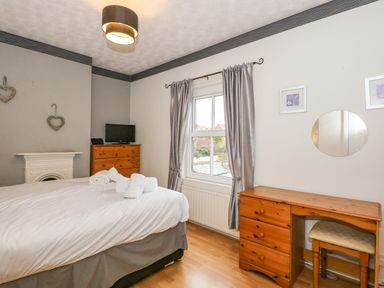 A bedroom with a bed with white linens a dresser with a small TV a wooden desk with a stool and window with curtains at 5 Melinda Cottages in East Runton near Cromer