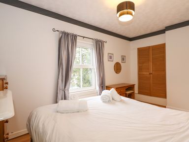 A bedroom with a bed with white linens and towels on it a window with curtains a wooden desk a wall mirror and framed pictures and a wooden wardrobe at 5 Melinda Cottages in East Runton near Cromer