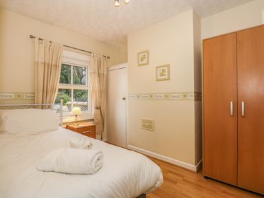 A bedroom with a bed with white linens and towels a bedside table with a lamp a window with curtains two framed pictures on the wall and a wooden wardrobe at 5 Melinda Cottages East Runton near Cromer
