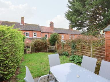 A garden with a patio table and chairs a wooden bench a wooden fence and brick houses in the background at 5 Melinda Cottages in East Runton near Cromer