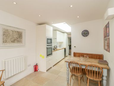 A kitchen with white cabinets a wooden dining table and chairs and a wall clock at Apple Tree Cottage in Shillingstone near Sturminster Newton