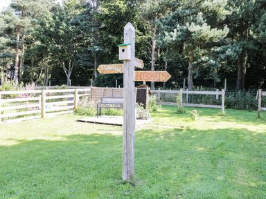 A wooden post with directional signs in a grassy fenced garden area surrounded by trees at Bunty's Place in Chatton near Wooler