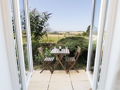 A patio with wooden table and two chairs with a bottle of wine and glasses overlooking fields at Buntys Place in Chatton near Wooler