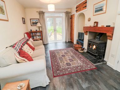 A living room with a beige sofa red and patterned cushions a wooden bookshelf a patterned rug a fireplace and glass doors leading outside at Buntys Place in Chatton near Wooler