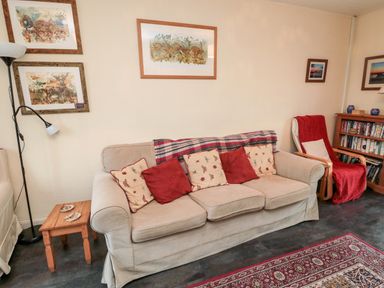A living room with a beige sofa with red and patterned cushions a wooden side table a standing lamp and framed artwork on walls at Bunty's Place in Chatton near Wooler
