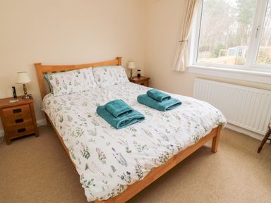 A bedroom with a double wooden bed with floral bedding two folded towels on the bed two wooden bedside tables with lamps a window with cream curtains and a radiator below it at Bunty's Place in Chatton near Wooler