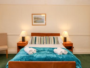 A bedroom with a double bed with striped pillows and rolled towels on a teal blanket two bedside tables with lamps a beige chair and a framed painting on the wall at Eden House in Edenhall near Penrith
