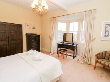 A bedroom with a bed topped with rolled towels a dark wood wardrobe and dresser a makeup table and chair by the bay windows and a striped armchair at Eden House Edenhall near Penrith