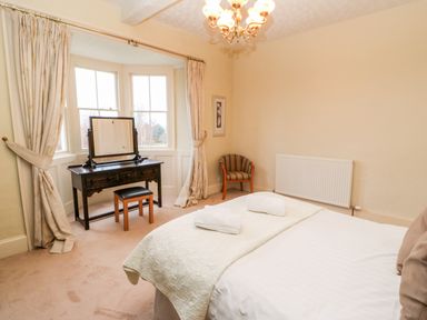 A bedroom with a bed, dressing table with mirror, a stool, a striped chair, windows with curtains, and a chandelier at Eden House in Edenhall near Penrith